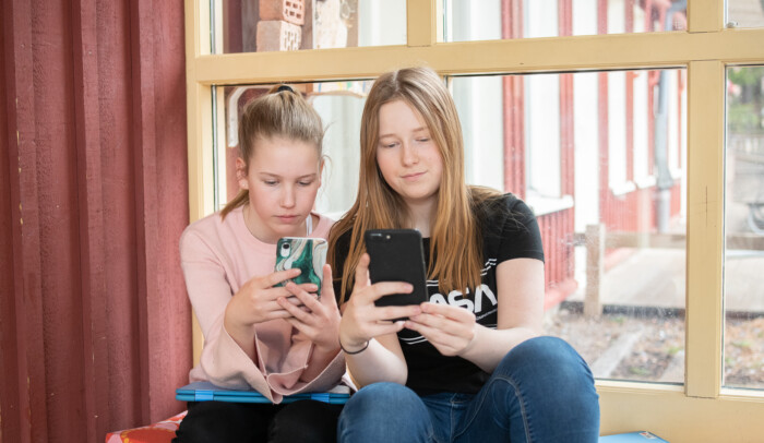 Två elever sitter vid ett fönster och tittar ner på sina mobiltelefoner